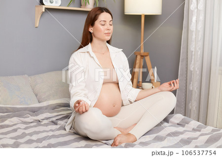 Meditating on maternity. Young caucasian pregnant woman practices yoga on bed at home in the morning Young beautiful pregnant woman sitting in lotus position good for fetus prenatal care. 106537754