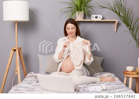 Smiling attractive pregnant brown haired caucasian woman sitting in bed having video call on laptop online conversation showing new little baby's clothing to notebook camera. Smiling attractive pregnant brown haired caucasian woman sitting in bed having video call on laptop online conversation showing new little baby's clothing to notebook camera. 106537842