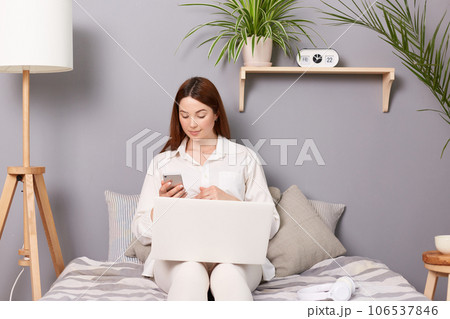 Pregnant woman with brown hair sitting on the bed in her bedroom taking a break, typing on her computer and glancing at her phone screen, future mother wearing white shirt. 106537846