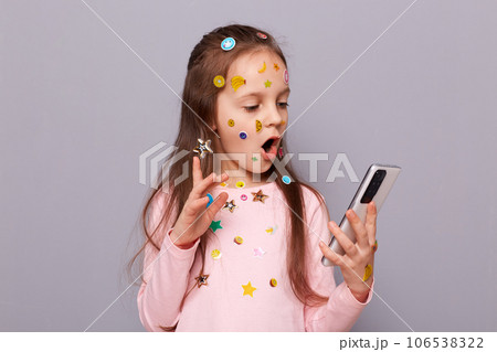 Shocked surprised little girl covered colorful kid's stickers standing isolated over gray background holding mobile phone in hands reading shocking news in internet. Shocked surprised little girl covered colorful kid's stickers standing isolated over gray background holding mobile phone in hands reading shocking news in internet. 106538322