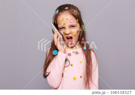 Funny crazy little girl covered colorful kid's stickers standing isolated over gray background talking on smartphone arguing with somebody grimacing making face. Funny crazy little girl covered colorful kid's stickers standing isolated over gray background talking on smartphone arguing with somebody grimacing making face. 106538327