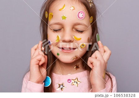 Hopeful little girl covered colorful kid's stickers standing isolated over gray background standing with closed eyes and crossed fingers making wish praying for good luck. Hopeful little girl covered colorful kid's stickers standing isolated over gray background standing with closed eyes and crossed fingers making wish praying for good luck. 106538328