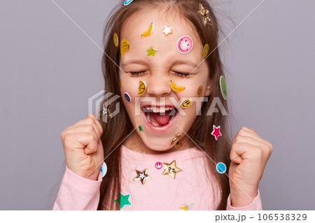 Extremely happy joyful little girl covered colorful kid's stickers standing isolated over gray background closing her eyes and clenched fists celebrating her success. Extremely happy joyful little girl covered colorful kid's stickers standing isolated over gray background closing her eyes and clenched fists celebrating her success. 106538329