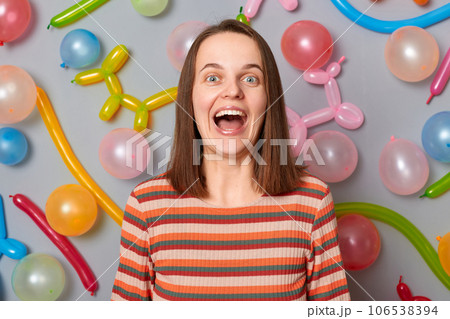 Amazed exited surprised woman wearing striped dress standing against gray wall with balloons decoration looking at camera with overjoyed expression. Amazed exited surprised woman wearing striped dress standing against gray wall with balloons decoration looking at camera with overjoyed expression. 106538394