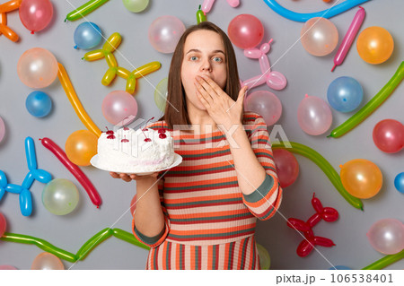 Amazed surprised woman with brown hair wearing striped dress holding cake covering mouth with hands sees shocked birthday present standing against gray wall decorated with colorful balloons. 106538401