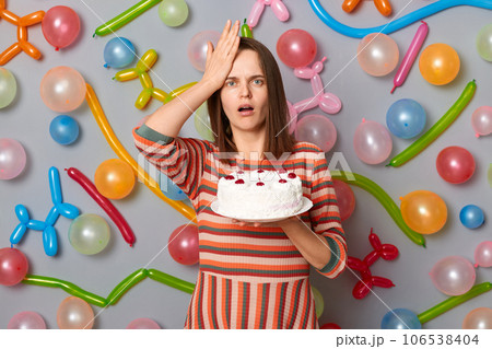 Shocked upset woman with brown hair wearing striped dress holding cake and looking at camera with open mouth, epic fail, standing against gray wall decorated with colorful balloons. Shocked upset woman with brown hair wearing striped dress holding cake and looking at camera with open mouth, epic fail, standing against gray wall decorated with colorful balloons. 106538404