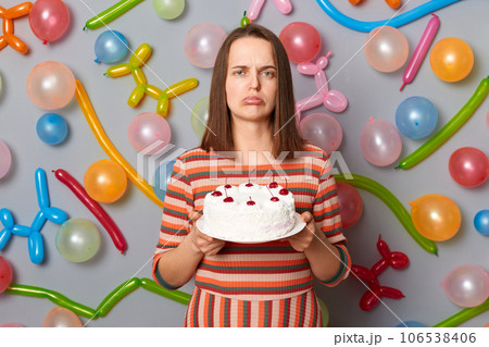 Sad disappointed woman with brown hair wearing striped dress holding cake she dislikes mixing order buying wrong dessert standing against gray wall decorated with colorful balloons. 106538406