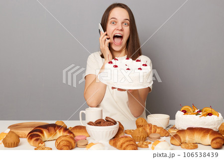 Amazed woman wearing white T-shirt isolated over gray background sitting at table with cake talking on cell phone screaming with happiness, keeps mouth open. 106538439