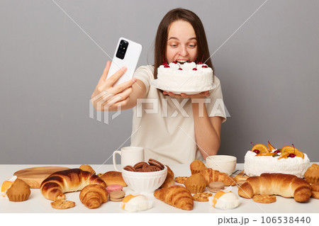 Funny woman blogger wearing white T-shirt isolated over gray background biting big cake biting dessert taking selfie creating content celebrating eating cheat meal. 106538440