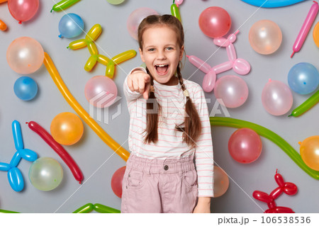Overjoyed excited little girl with pigtails standing against gray wall with balloons decoration pointing to camera with amazement screaming celebration. 106538536
