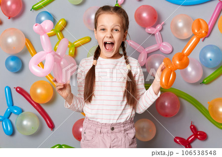 Extremely excited festive little girl  with pigtails standing against gray decorated colorful wall holding balloons in hands screaming hurray celebrating birthday. 106538548