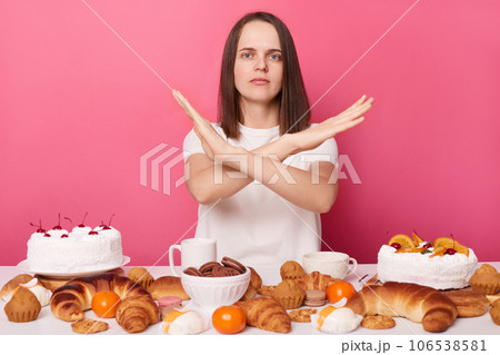 Strict brown haired woman in white t shirt sitting at table with pastry showing no gesture crossing hands stop eating sweets isolated over pink background. Strict brown haired woman in white t shirt sitting at table with pastry showing no gesture crossing hands stop eating sweets isolated over pink background. 106538581