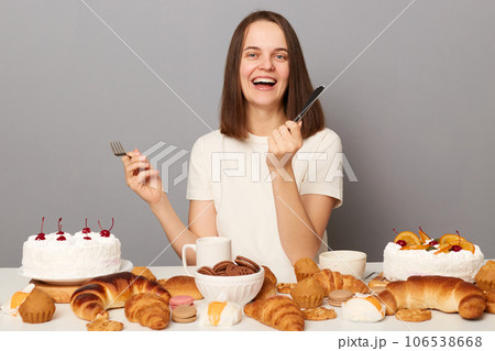 Extremely happy positive woman wearing white T-shirt isolated over gray background sitting at festive table among bakery sugary desserts holding fork and knife. Extremely happy positive woman wearing white T-shirt isolated over gray background sitting at festive table among bakery sugary desserts holding fork and knife. 106538668