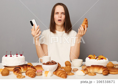 Shocked scared woman wearing white T-shirt isolated over gray background sitting at table with desserts holding mobile phone and croissant looking at camera with frightened expression. 106538671