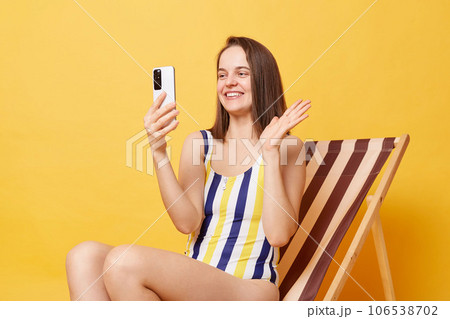 Resort blog. Smiling woman blogger wearing striped swimming suit sitting on deck chair isolated over yellow background, broadcasting livestream from vacation, having video call, saying hi. Resort blog. Smiling woman blogger wearing striped swimming suit sitting on deck chair isolated over yellow background, broadcasting livestream from vacation, having video call, saying hi. 106538702