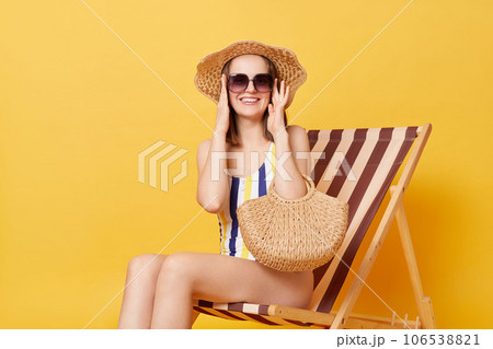 Smiling relaxed woman wearing striped swimming suit sunglasses and hat sitting on deck chair isolated over yellow background relaxing on beach enjoying vacation. Smiling relaxed woman wearing striped swimming suit sunglasses and hat sitting on deck chair isolated over yellow background relaxing on beach enjoying vacation. 106538821