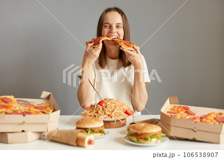 Hungry funny woman with brown hair wearing white T-shirt sitting at table among fast food isolated over gray background holding two slices of pizza eating junk food. Hungry funny woman with brown hair wearing white T-shirt sitting at table among fast food isolated over gray background holding two slices of pizza eating junk food. 106538907