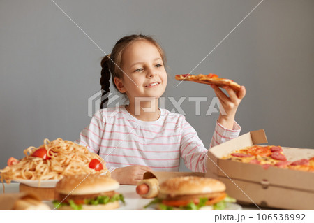 Enjoyable smiling beautiful little girl with braids sitting at table with junk food isolated over gray background holding big slice of pizza being reading to eat uhealthy dish. 106538992