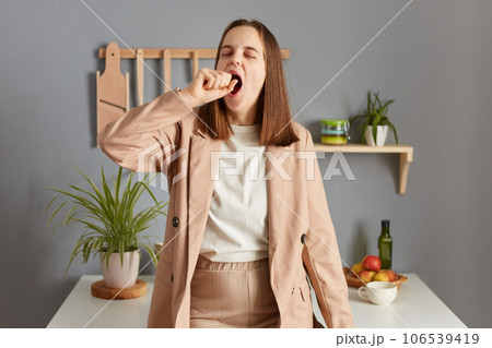 Sleepy worker. Early awaking. Yawning woman with brown hair wearing beige jacket standing in home kitchen interior covering mouth with palm being sleepless. 106539419