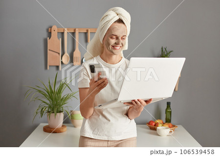 Charming adorable young woman posing in kitchen with towel on head doling her freelance tasks while taking care of her skin making beauty treatment procedures. Charming adorable young woman posing in kitchen with towel on head doling her freelance tasks while taking care of her skin making beauty treatment procedures. 106539458