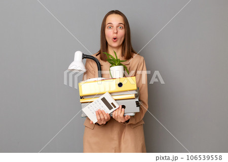 Amazed surprised woman holding cardboard box with her things lost job, posing isolated on gray background looking at camera with open mouth. Amazed surprised woman holding cardboard box with her things lost job, posing isolated on gray background looking at camera with open mouth. 106539558