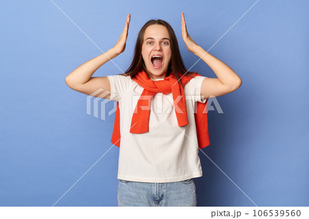 Positive cheerful surprised woman wearing white t-shirt and jumper over neck standing isolated over blue background raised arms being amazed and excited hearing great news. Positive cheerful surprised woman wearing white t-shirt and jumper over neck standing isolated over blue background raised arms being amazed and excited hearing great news. 106539560