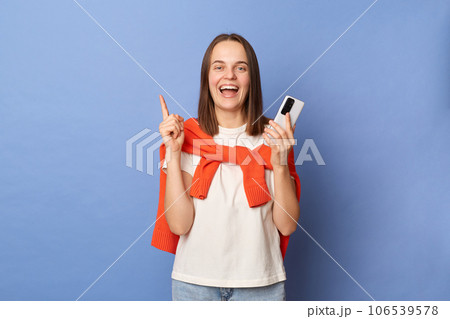 Excited inspired dark haired woman wearing orange jumper over shoulders using mobile phone raising finger pointing at copy space standing isolated over blue background. Excited inspired dark haired woman wearing orange jumper over shoulders using mobile phone raising finger pointing at copy space standing isolated over blue background. 106539578