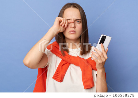 Tired dark haired woman wearing orange jumper over shoulders standing isolated over blue background using mobile phone long hours feeling pain in her eyes. Tired dark haired woman wearing orange jumper over shoulders standing isolated over blue background using mobile phone long hours feeling pain in her eyes. 106539579