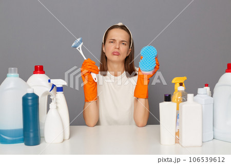 Sad upset unhappy woman posing at workplace with cleaning detergents holding sponge and brush crying looking at camera with pout lips isolated over gray background. 106539612