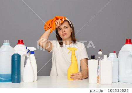 Exhausted woman posing at workplace with cleaning detergents isolated over gray background working long hours having headache looking at camera with tired expression. Exhausted woman posing at workplace with cleaning detergents isolated over gray background working long hours having headache looking at camera with tired expression. 106539625