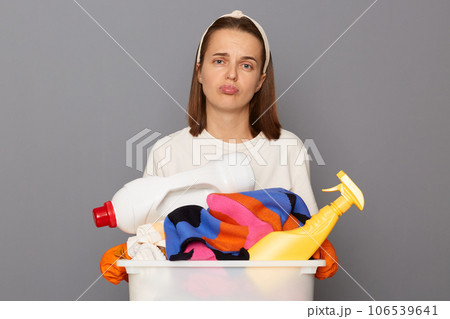 Exhausted woman wearing whote t-shirt with laundry bowl and detergent isolated over gray background feeling bored and unhappy needs rest looking at camera with sad expression. Exhausted woman wearing whote t-shirt with laundry bowl and detergent isolated over gray background feeling bored and unhappy needs rest looking at camera with sad expression. 106539641