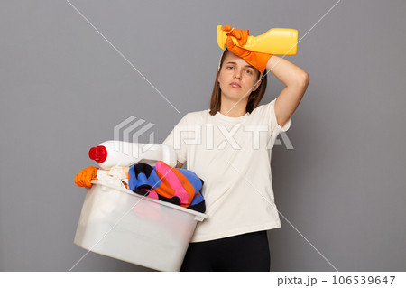 Housewife takes care of clothing. Domestic appliances aid in housework. Tired exhaused woman wearing whote t-shirt with laundry bowl and detergent isolated over gray background Housewife takes care of clothing. Domestic appliances aid in housework. Tired exhaused woman wearing whote t-shirt with laundry bowl and detergent isolated over gray background 106539647