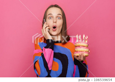 Amazed news on birthday. Shocked astonished female in colorful sweater holding delicious sweets with candles talking phone with excited look, saying wow isolated over pink background. 106539654