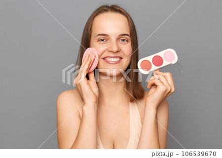 Skin care and beauty. Daily makeup. Smiling attractive young woman with brown hair holding sponge and powder posing isolated over gray background. Skin care and beauty. Daily makeup. Smiling attractive young woman with brown hair holding sponge and powder posing isolated over gray background. 106539670