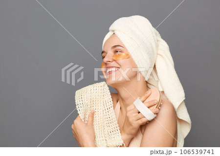 Dry massage. Shower beauty procedures. Delighted smiling young woman wrapped head in towel with patches under eyes doing skin care procedures posing isolated over gray background. 106539741