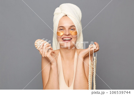 Caring for body. Skin care treatment. Shower routine. Cheerful satisfied young woman wrapped head in towel with patches under eyes posing isolated over gray background. 106539744