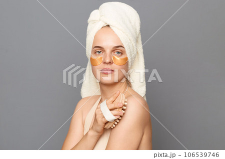 Beauty portrait of attractive calm relaxed young woman wrapped head in towel with patches under eyes dry brushing her arm, taking care of her body posing isolated over gray background. 106539746