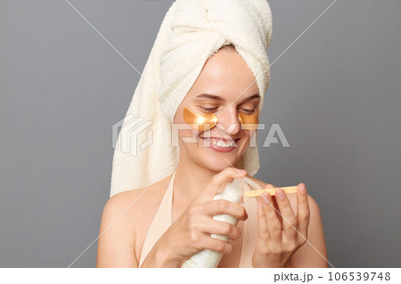 Cosmetic procedures. Skin care. Happy joyful young woman wrapped head in towel with patches under eyes using lotion to take care of face skin posing isolated over gray background. Cosmetic procedures. Skin care. Happy joyful young woman wrapped head in towel with patches under eyes using lotion to take care of face skin posing isolated over gray background. 106539748