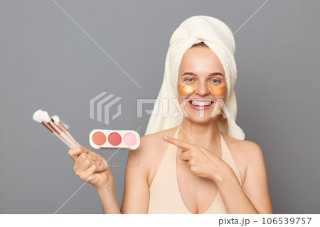 Beauty promotion. Makeup products. Happy woman model with patches under eyes wrapped in towel pointing at powder and brushes advertising isolated over gray background 106539757