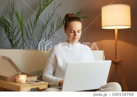 Remote work. Freelance job. Concentrated focused woman with bun hairstyle in white sweater looking at laptop screen typing keyboard working sitting on sofa at home. Remote work. Freelance job. Concentrated focused woman with bun hairstyle in white sweater looking at laptop screen typing keyboard working sitting on sofa at home. 106539769