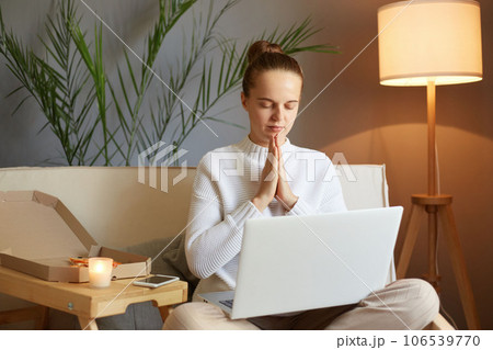 Relaxed woman with bun hairstyle in white sweater posing in front of laptop screen sitting on sofa at home meditating practicing yoga while having break at work. Relaxed woman with bun hairstyle in white sweater posing in front of laptop screen sitting on sofa at home meditating practicing yoga while having break at work. 106539770