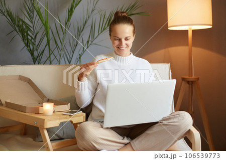 Relaxed happy smiling woman with bun hairstyle in white sweater looking at laptop screen and having snack eating pizza while watching film sitting on sofa at home. Relaxed happy smiling woman with bun hairstyle in white sweater looking at laptop screen and having snack eating pizza while watching film sitting on sofa at home. 106539773