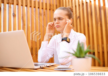 Tired concentrated woman wearing white shirt sitting in outdoor cafe working on laptop massaging templas has headache thinking about new job tasks. 106539833