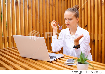 Uncertain puzzled woman wearing white shirt sitting in outdoor cafe working on laptop looking at computer monitor with puzzled expression. 106539835