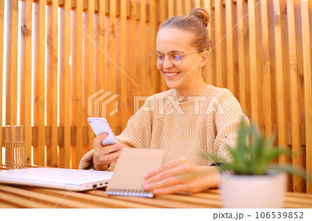 Carefree woman wearing beige sweater sitting in outdoor cafe working on laptop using phone and paper planer planning her work enjoying freelance work. 106539852