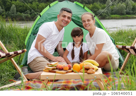 Happy family sitting near tent on the ground having picnic together enjoying tasty fruit and sandwiches, spending happy weekend in open air. Happy family sitting near tent on the ground having picnic together enjoying tasty fruit and sandwiches, spending happy weekend in open air. 106540051