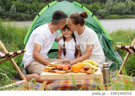 Camping near the river in nature park. Family at vacation. Couple with daughter resting. Parents kiss=sing daughter. People enjoying vacation. Camping near the river in nature park. Family at vacation. Couple with daughter resting. Parents kiss=sing daughter. People enjoying vacation. 106540058