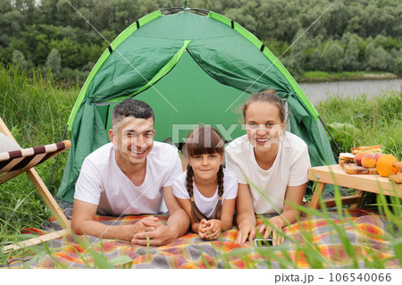 Nature vacation. Family trip. Camping trip, Mother daughter father lying on grass and blanket looking at camera, enjoying resting near the lake or river. 106540066