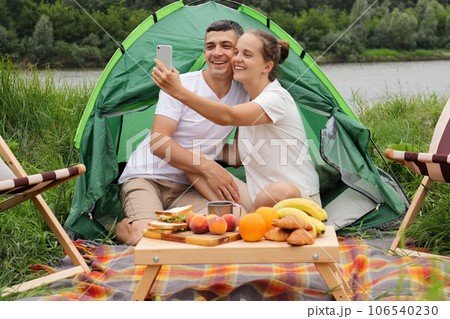 Happy woman and man wearing casual clothing sitting at tent near the river making selfie on smart phone enjoying resting in nature, Happy woman and man wearing casual clothing sitting at tent near the river making selfie on smart phone enjoying resting in nature, 106540230