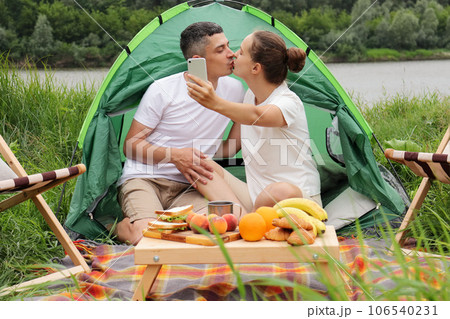 happy time in nature. Camping at riverside. Romantic woman and man wearing casual clothing sitting at tent near the river, couple kissing and making selfie. 106540231